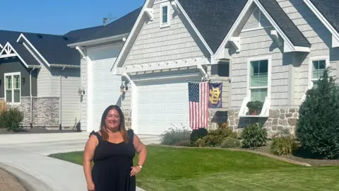 Christine Carlson, una mujer que usa un vestido negro, posa en un día soleado frente a una casa familiar. La casa está pintada de blanco y tiene un césped delantero herbáceo, y tiene una bandera estadounidense colgada en el frente.