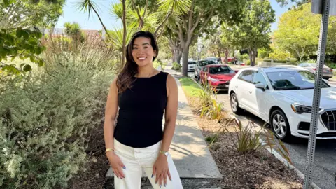 Aileen Barameda Una mujer que lleva una camisa negra y pantalones blancos posa en la acera de una calle residencial bordeada de árboles al lado de un automóvil blanco estacionado.