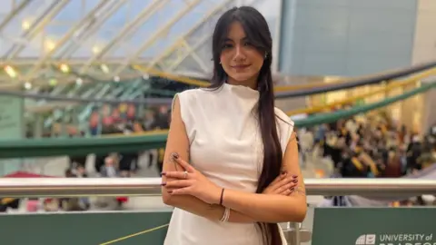 Sanskrity Baraili Sanskrity se encuentra frente a una sala concurrida. Lleva un vestido blanco y mira fijamente a la cámara.